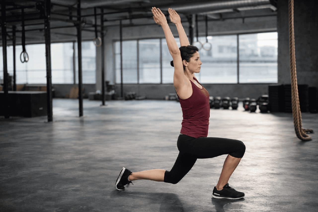 En kvinna gör ett utfall med armarna sträckta över huvudet i ett gym