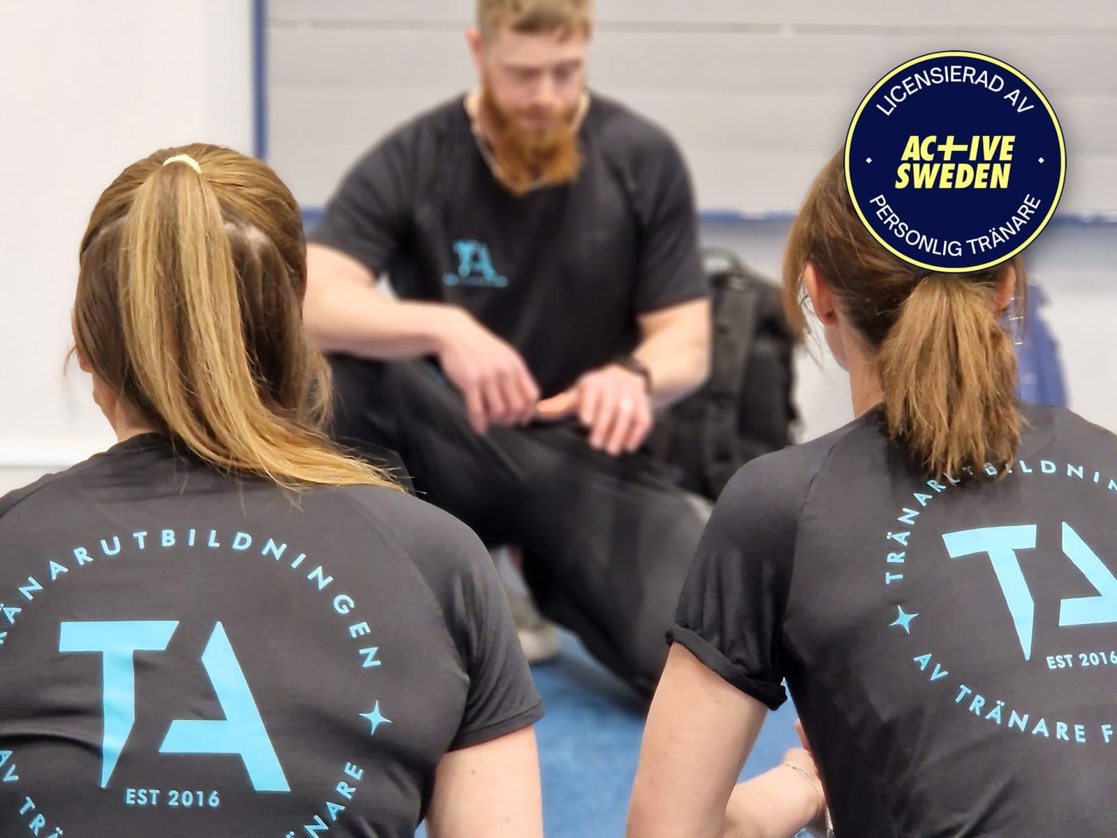 Två studenter i TA-tröjor lyssnar på läraren i lektionssalen, med Active Swedens sigill Licensierad av Active Sweden, personlig tränare