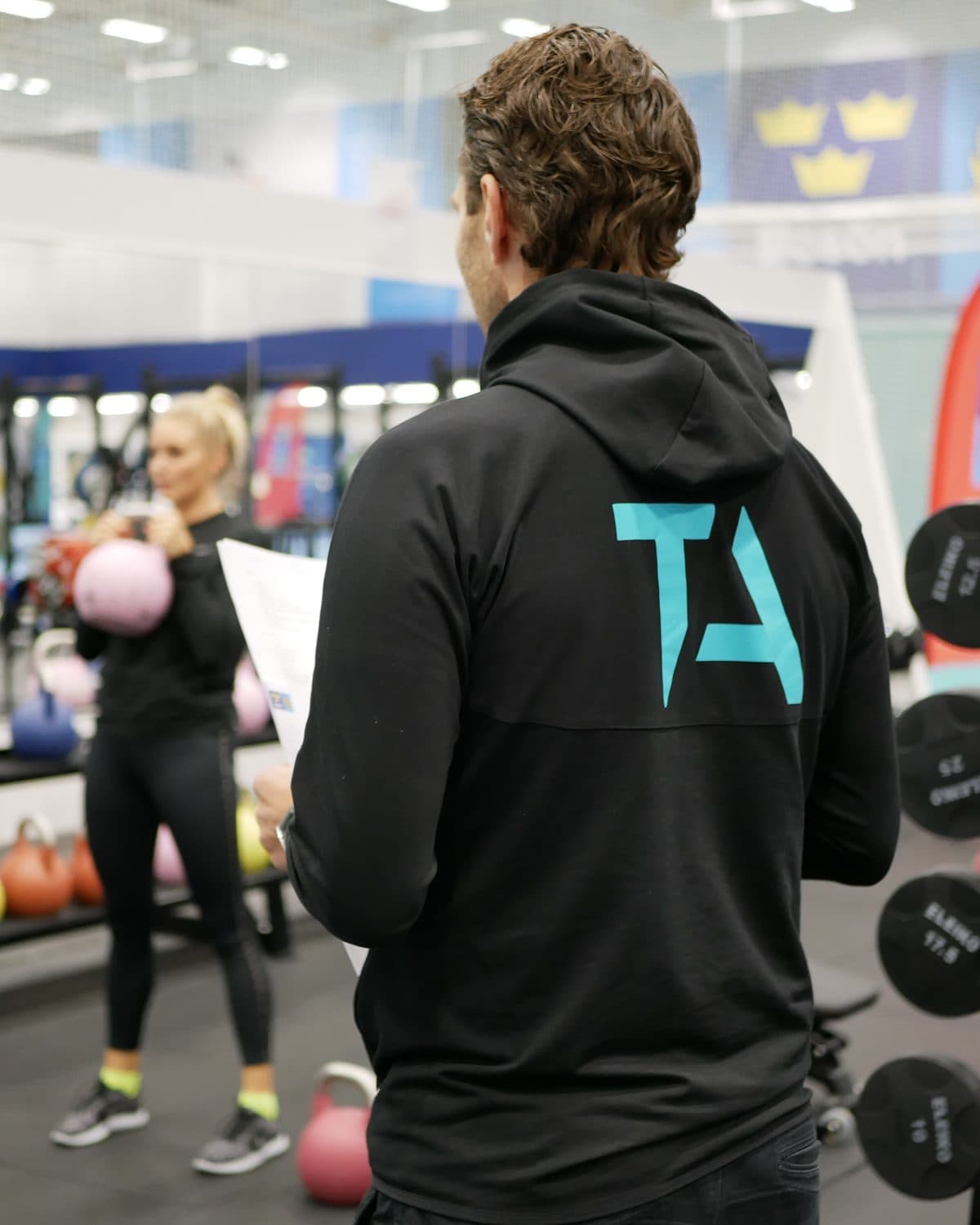 En lärare i TA-hoodie följer en kund som tränar med kettlebell i Framgångshallen på Bosön