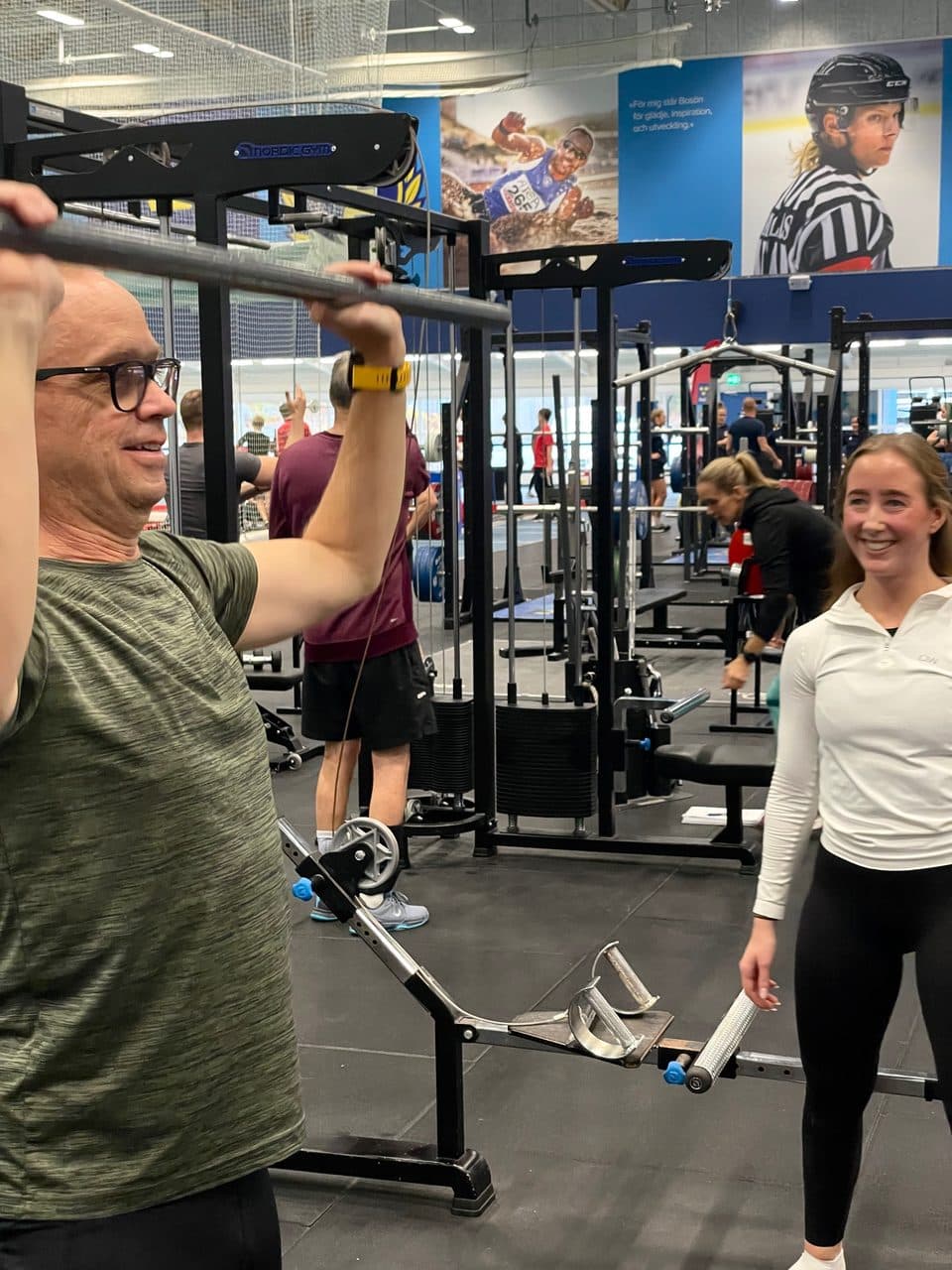 En man lyfter en stång medan en kvinna står bredvid och ler i gymmet.